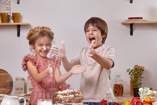 Pretty boy dressed in a light t-shirt and jeans and a little girl with a braid in her hair, wearing in a pink dress are making a cake at a kitchen, against a white wall with shelves on it. Children smeared their hands with powdered sugar, laughing and clapping it.