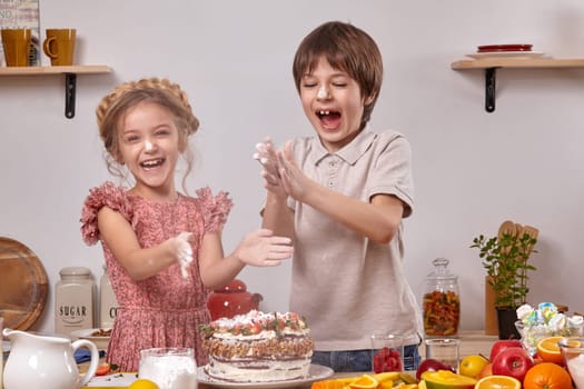 Funny boy dressed in a light t-shirt and jeans and a little girl with a braid in her hair, wearing in a pink dress are making a cake at a kitchen, against a white wall with shelves on it. They smeared their hands with powdered sugar, laughing and clapping it.
