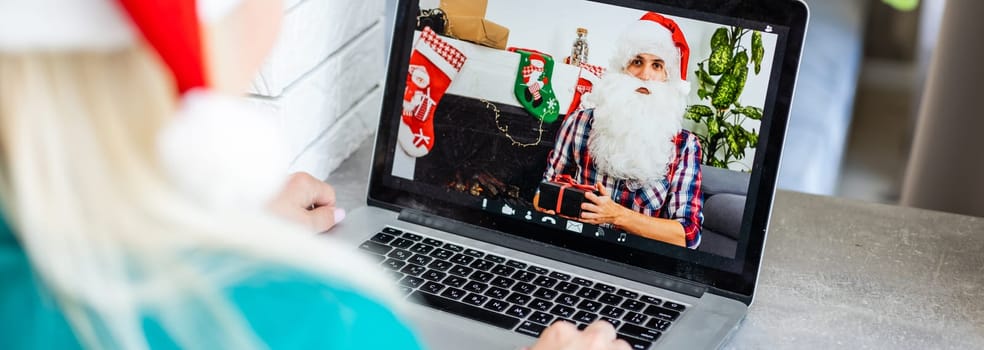 Smiling girl wearing Santa hat having video calling family by webcam. Woman with laptop sitting on kitchen at home using virtual meeting chat on holidays. Happy Christmas and New Year new normal