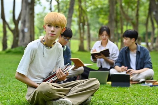 Asian male student sitting on green grass in campus with friends sitting on background. Education and lifestyle concept.