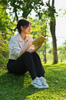 Calm female university student reading book, relaxing on campus lawn. Education, learning and lifestyle concept.