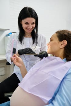 Beautiful pregnant woman, patient at dental appointment, sitting in dentist's chair, smiling in mirror, discussing with the doctor the teeth shade and color for bleaching procedure in dentistry clinic
