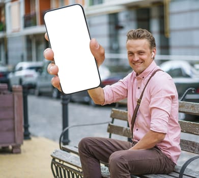 Man sitting on city bench on street, holding mobile phone with white screen display that offers copy space. Integration of technology and urban lifestyle, man engages with mobile device in public. . High quality photo