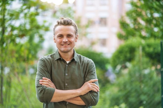 Blond, smiling, and handsome Caucasian man posing for camera in city park. Man's charming smile and confident demeanor radiate happiness and joy. Natural beauty of the outdoor setting, with lush greenery and the urban cityscape in background. High quality photo