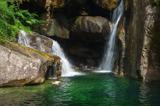 An unrecognizable person swims through crystal clear emerald green waters towards a hidden cave behind a falling cascade, a secret place unknown to many, but desired for social recognition.