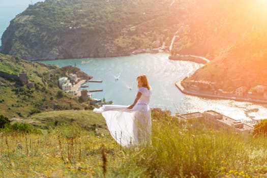 Happy woman in a white dress and hat stands on a rocky cliff above the sea, with the beautiful silhouette of hills in thick fog in the background