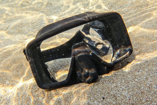 Underwater photo - sun shining to rubber black diving mask on sand in shallow water.