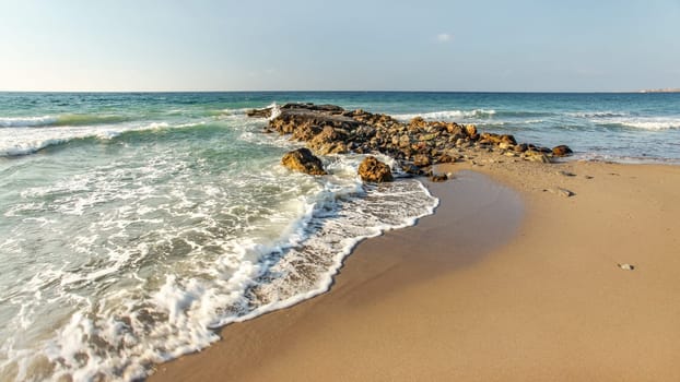 Afternoon sun shines on wild beach, some small waves hitting little rocks in background, empty clean sand spot (space for text) on right side.