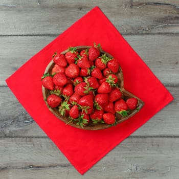 Top down view, carved wooden bowl with strawberries on red tablecloth and gray wood desk.