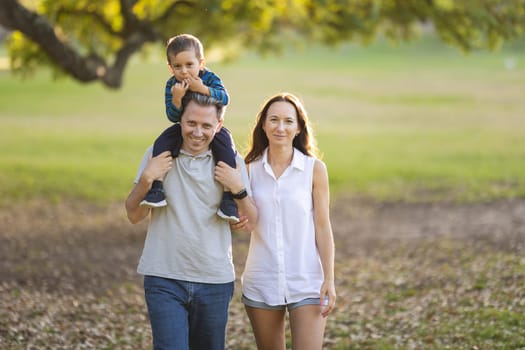 Cute white smiling family in the park - little son on the shoulders of his father and his wife standing by - looking in the camera. Mid shot