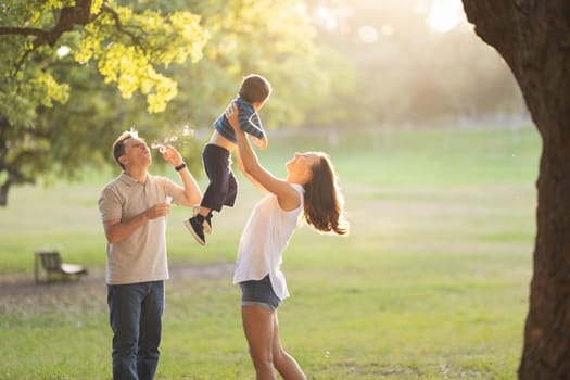 Happy white family spending time in the park - mom raising her son in the air while father blowing soap bubbles. Mid shot