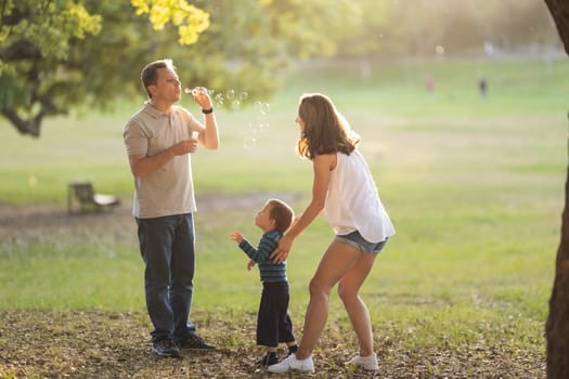 Happy white family spending time in the park - father blowing soap bubbles. Mid shot
