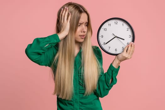 Confused woman with anxiety checking time on clock, running late to work, being in delay, deadline. Young teen girl looking at hour minutes, worrying to be punctual isolated on pink studio background
