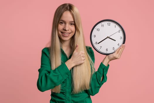 It is your time. Caucasian smiling woman showing time on wall office clock, ok, thumb up, approve, pointing finger at camera, hurry up, approve. Young teen girl isolated on pink studio background