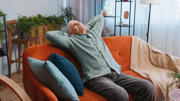 Tired grandfather man enjoy relaxing on home sofa in room. Happy senior pensioner sits on couch resting napping after hard working day, closed his eyes, put his hands behind his head, stress free