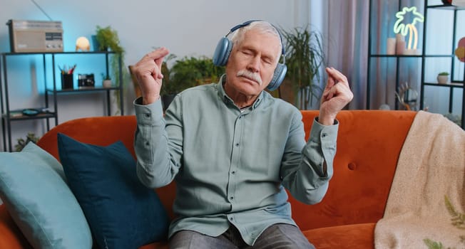 Keep calm down, relax, inner balance. Grandfather senior man breathes deeply with mudra gesture, eyes closed, meditating with concentrated thoughts, peaceful mind at home. Elderly guy sitting on couch