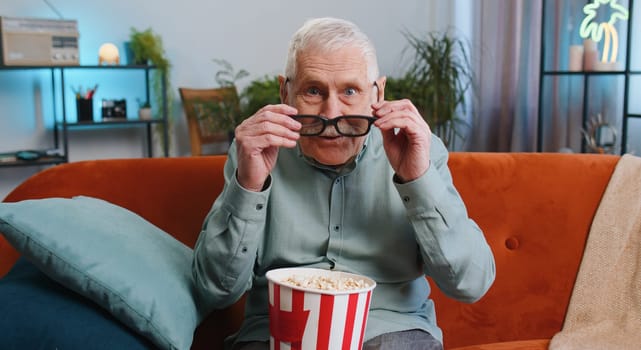 Senior grandfather man in 3D glasses on sofa eating popcorn snacks and watching interesting TV serial sport game, film, online social media movie content at home apartment. Guy enjoying entertainment