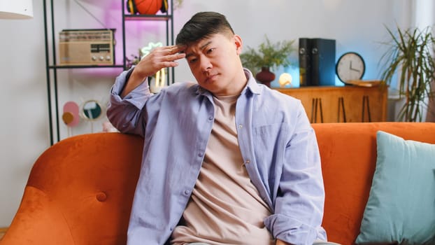 Sad lonely man sitting at home room looks pensive thinks over life concerns or unrequited love suffers from unfair situation. Chinese alone guy problem, break up, depressed feeling bad annoyed burnout