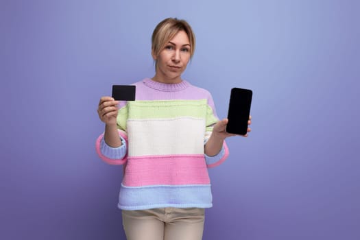blond young woman in casual outfit showing credit card and smartphone with mockup on purple background with copy space.