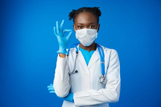 Portrait happy African American female doctor showing ok sign in blue studio, close up