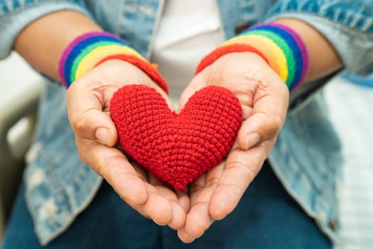 Asian lady wearing rainbow flag wristbands and hold red heart, symbol of LGBT pride month celebrate annual in June social of gay, lesbian, bisexual, transgender, human rights.