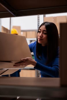 Goods supply chain manager doing inventory in warehouse while checking product box on shelf. Asian woman storehouse worker taking freight carton from rack in storage room
