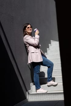 Stylish Beautiful Brunette Woman Drinks Coffee From Paper Cup, Standing On Stairs Near Office Building Outside, Summer Sunny Day. Taking A Break. Lifestyle Mature Female. Vertical Plane.
