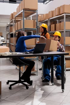 Warehouse manager working on laptop and taking parcel from employee in wheelchair. Post office storehouse asian colleagues cooperating and managing packages distribution process