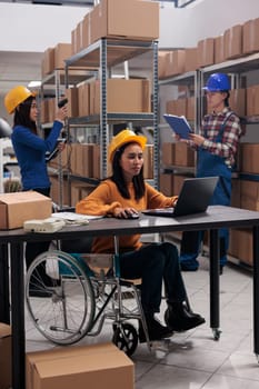 Order fulfillment warehouse worker in wheelchair doing goods stock management on laptop. Storehouse employee with disability tracking parcel online in disability inclusive workplace