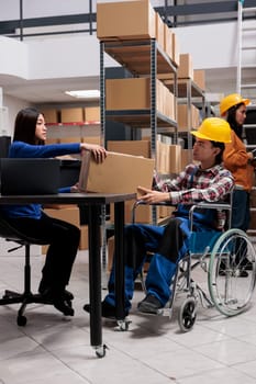 Warehouse employees managing received package in storage room. Asian man with disability and woman distribution managers doing logistics operations while holding parcel in storehouse