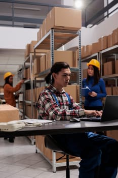 Logistics manager planning goods supply on laptop in freight warehouse. Young asian shipment service employee typing on computer and checking inventory management system in storehouse