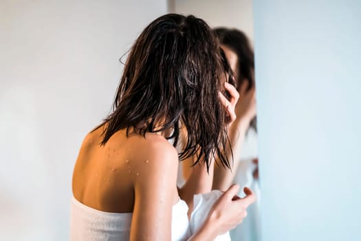 After a shower and hygienic procedures, a young pretty girl, wrapped in a towel, looks at herself in the mirror, applies a mask on her hair, does skin care at home.