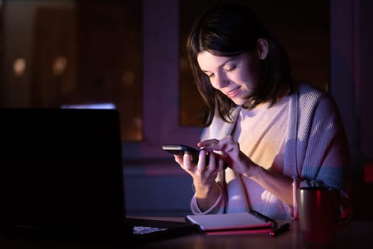 A smiling girl chats, communicates with colleagues and successfully develops her business at home at night, a young woman works on a laptop and holds a mobile phone in her hands.