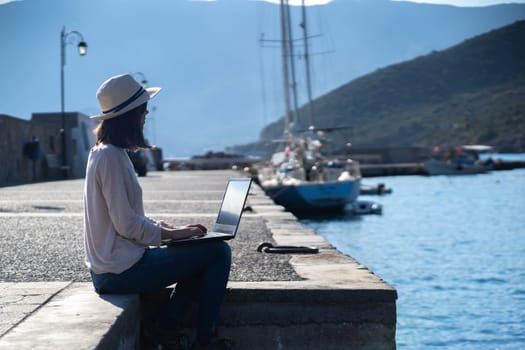 A young girl in a hat is sitting on the pier and working, typing on a laptop keyboard on a sunny day against a beautiful background of a seascape with moored yachts in the bay. Woman works and travels