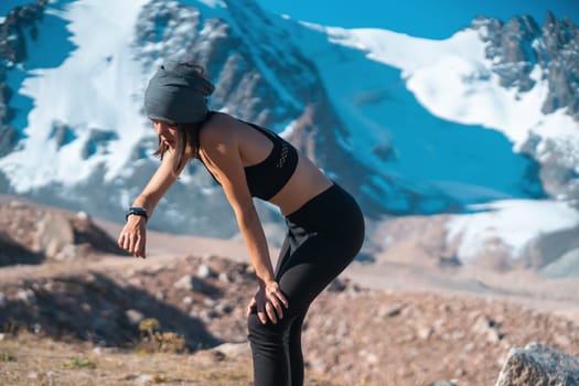 A young athletic girl looks at a smartwatch on her hand, checks her pulse, takes a break after jogging on trails in the snowy mountains. Woman runs outdoors in a mountainous beautiful area.