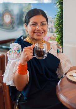 indigenous woman in traditional clothing, sitting in a cafeteria looking at the camera toasting very happy with a coffee in her hand