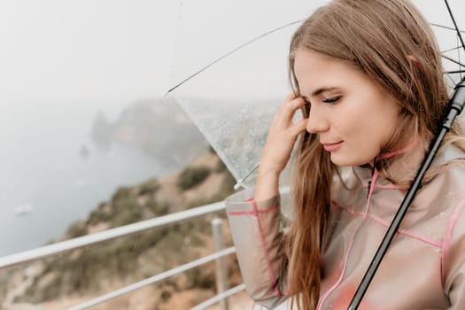 Woman rain park. Happy woman portrait wearing a raincoat with transparent umbrella outdoors on rainy day in park near sea. Girl on the nature on rainy overcast day