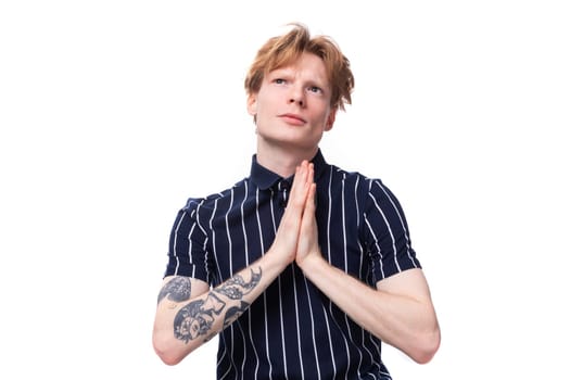 dreamy 25 year old blond man in striped polo praying on white background.