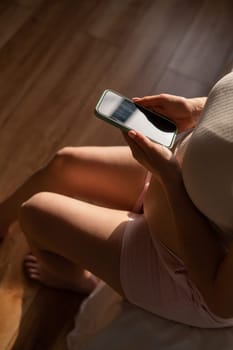 A pregnant woman uses a smartphone while sitting on the bed. Close-up of a pregnant woman's belly