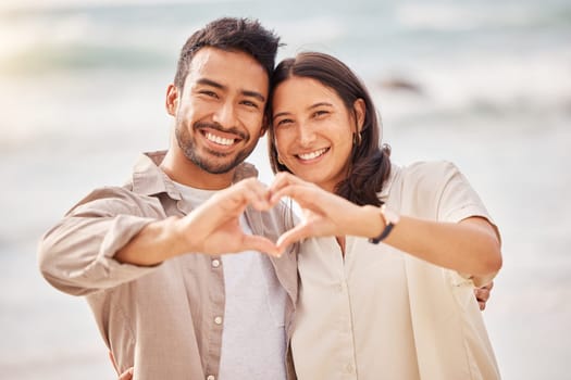 She completes me. Couple, portrait and love sign with hands at the ocean for travel with sunshine with a smile. Beach, together and heart with hand and smile in the summer for a weekend with romance