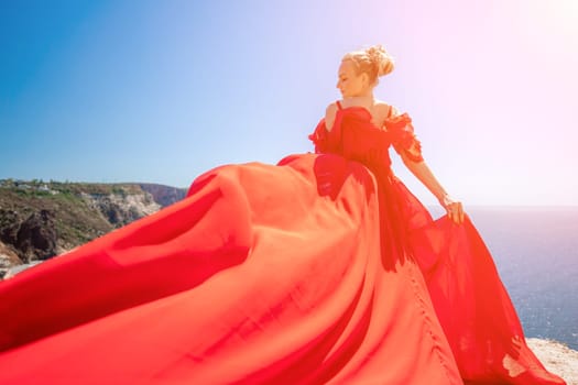 woman sea red dress. Blonde with long hair on a sunny seashore in a red flowing dress, back view, silk fabric waving in the wind. Against the backdrop of the blue sky and mountains on the seashore