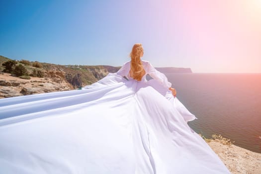 woman sea white dress. Blonde with long hair on a sunny seashore in a white flowing dress, rear view, silk fabric waving in the wind. Against the backdrop of the blue sky and mountains on the seashore