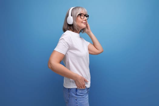 fashionable energetic woman 60s with gray hair listens to music in wireless headphones on studio background with copy space.