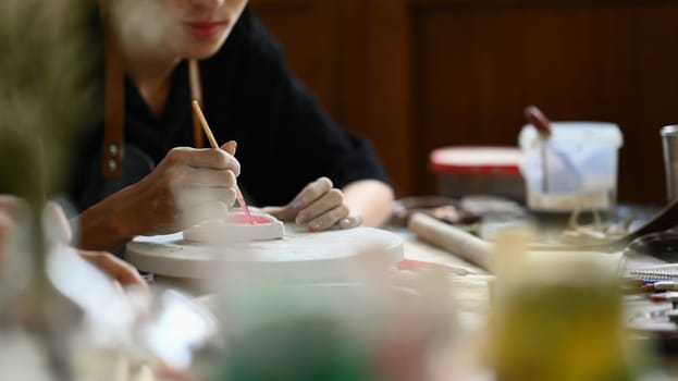 Cropped shot of clam young man in apron painting ceramic plate at workshop. Indoors lifestyle activity, handicraft, hobbies concept.