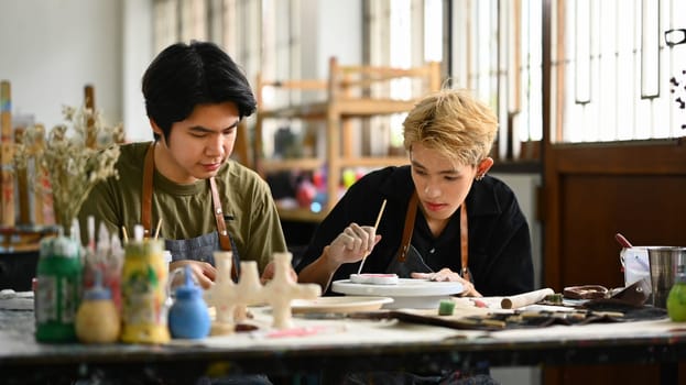 Smiling male potter teaching student man creating new handmade earthenware items in pottery workshop.