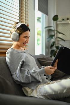 Casual young woman wearing headphone and using digital tablet on sofa in bright living room.