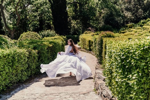 Brunette runs white dress park. A beautiful woman with long brown hair and a long white dress runs along the path along the beautiful bushes in the park, rear view.