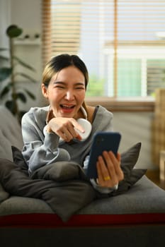 Happy young woman enjoying online virtual chat video call with friends via mobile phone while lying on couch at home.