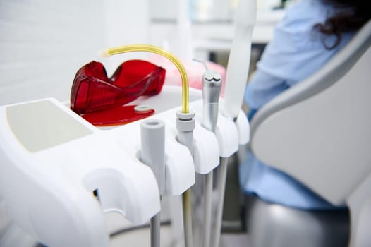 Selective focus. Close-up dental equipment with drill, saliva ejector over blurred background of patient in dentist's chair, getting teeth treatment in dentistry clinic