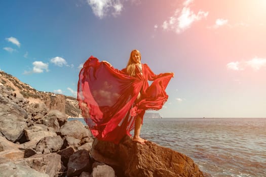 Red dress sea woman. A blonde with flowing hair in a long flowing red dress stands on a rock near the sea. Travel concept, photo session at sea.